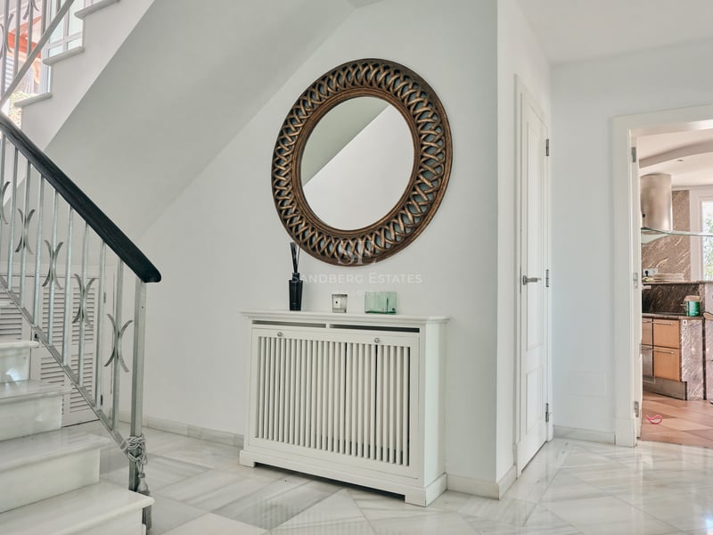 Entrée lumineuse avec escalier en marbre, cache-radiateur blanc et grand miroir circulaire orné de dorures.