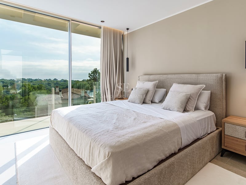 Chambre parentale moderne avec baies vitrées du sol au plafond donnant sur un paysage verdoyant et grand lit neutre.
