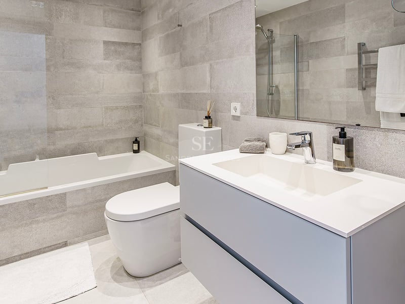 Salle de bain contemporaine avec carrelage gris, baignoire blanche, toilettes et meuble vasque moderne.