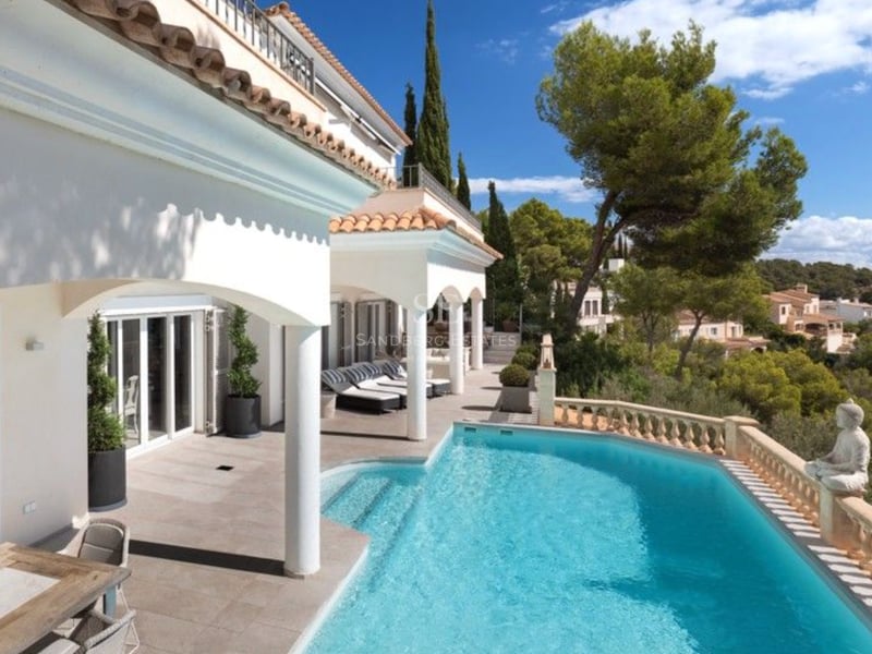 Vue en hauteur d'une villa blanche avec des arches, une terrasse en pierre, une piscine turquoise et des pins.