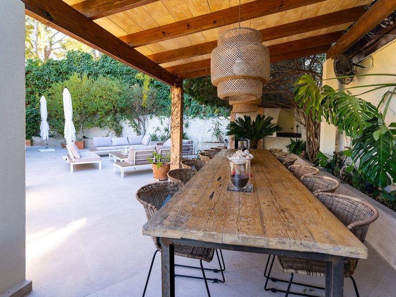 Longue table à manger en bois sous une pergola avec chaises en osier et suspension près d'un salon.