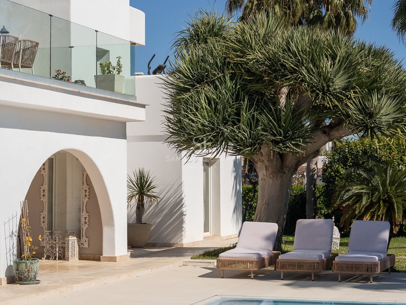 Piscine étincelante à côté d'une villa blanche avec des chaises longues et des palmiers sous un ciel bleu.