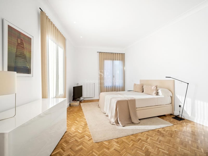 Chambre lumineuse avec parquet en chevrons, grand lit, rideaux beiges et lampe design.