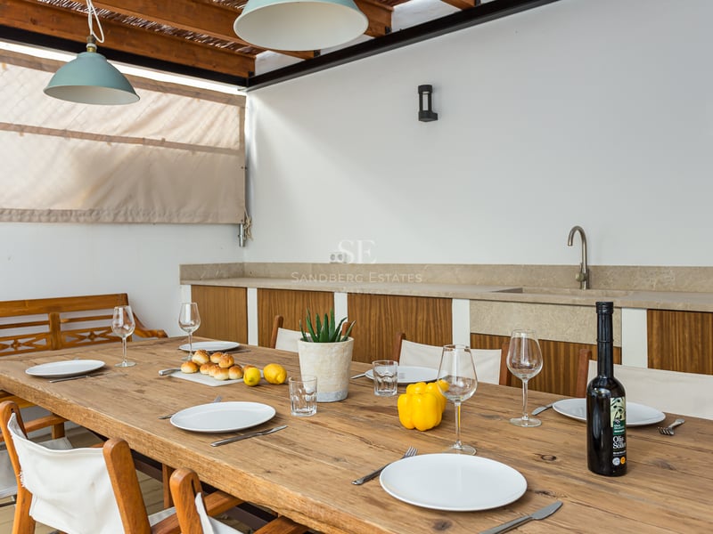 Longue table en bois dressée pour des invités sur une terrasse couverte avec cuisine extérieure.