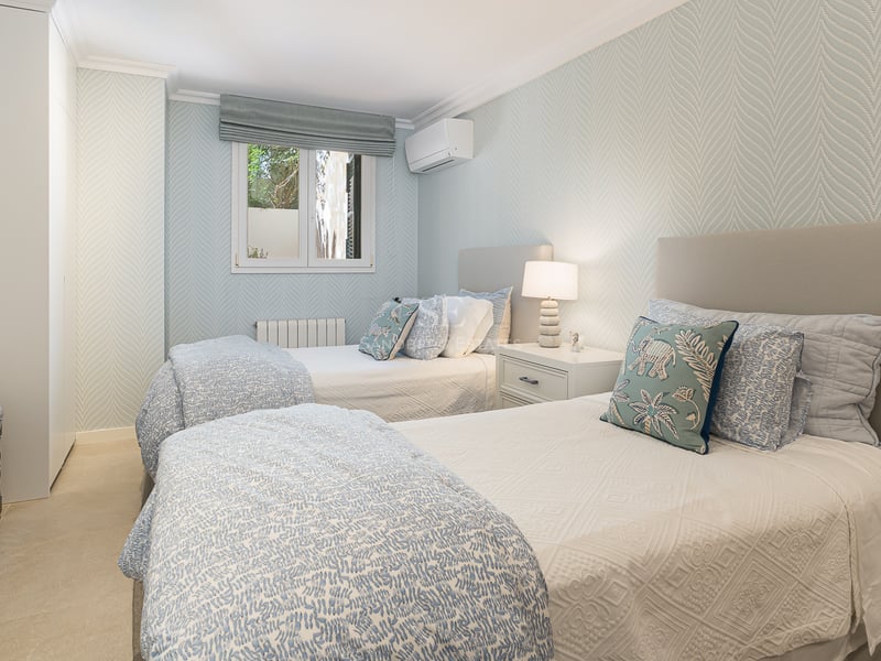 Chambre lumineuse avec deux lits simples, papier peint bleu clair, armoires encastrées et sol en pierre naturelle.