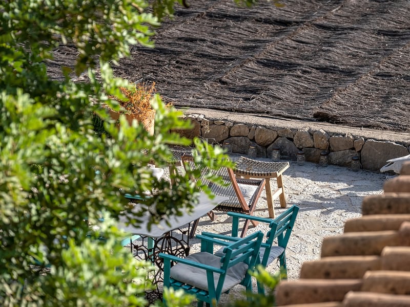 Terrasse en pierre avec chaises en bois bleu sarcelle et toit en chaume, vue à travers la verdure.