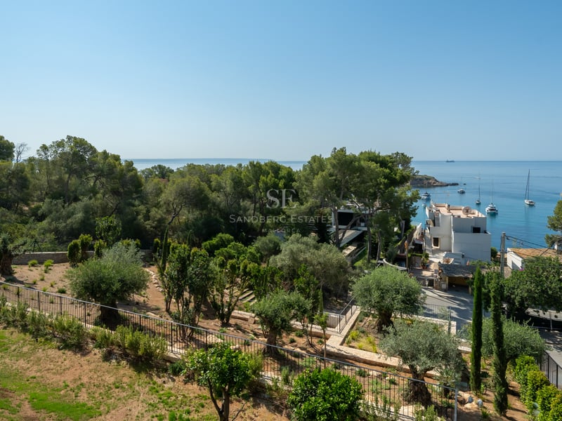 Vue d'un jardin méditerranéen avec oliviers surplombant une baie bleue avec voiliers et maisons blanches.