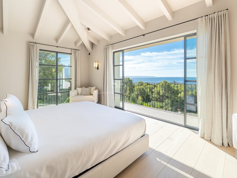 Chambre principale lumineuse avec poutres blanches, parquet en chêne et balcon offrant une vue sur la mer et la forêt.