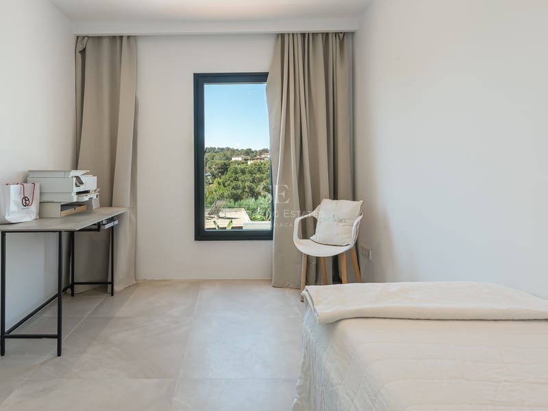 Chambre lumineuse avec lit blanc, bureau minimaliste et fenêtre offrant une vue sur les collines.