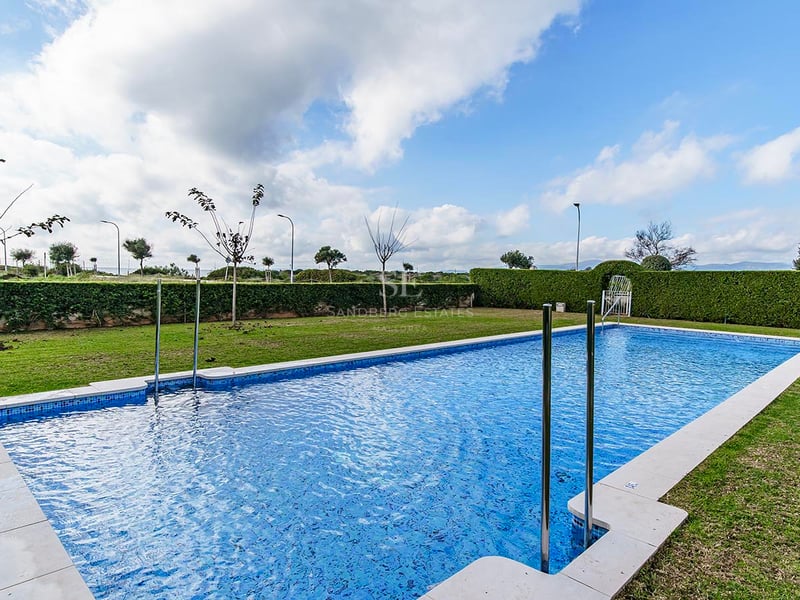 Grande piscine rectangulaire entourée d'une pelouse verte et de haies taillées sous un ciel nuageux.