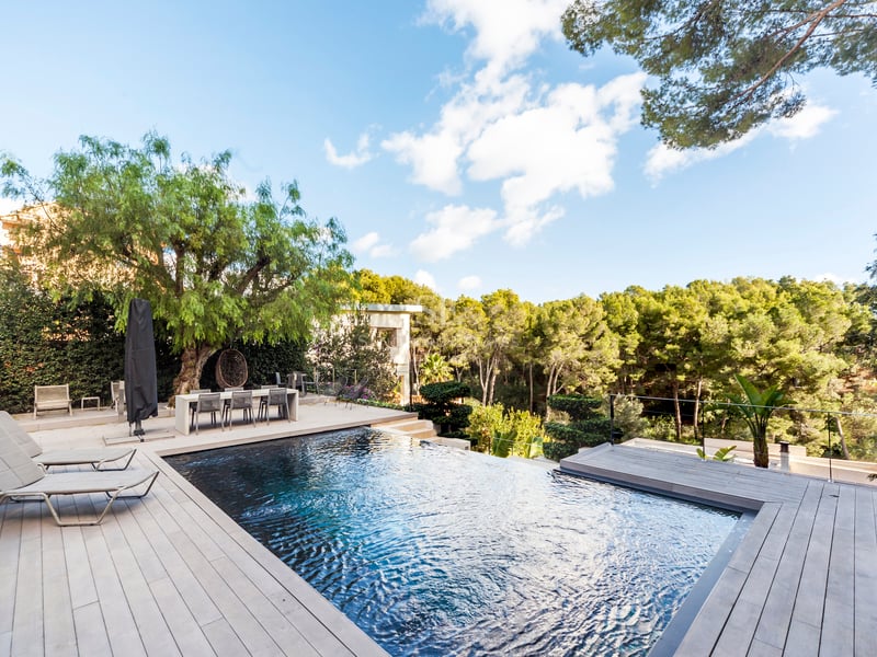 Piscine à débordement de luxe entourée d'une terrasse en bois et d'arbres verdoyants sous un ciel bleu clair.