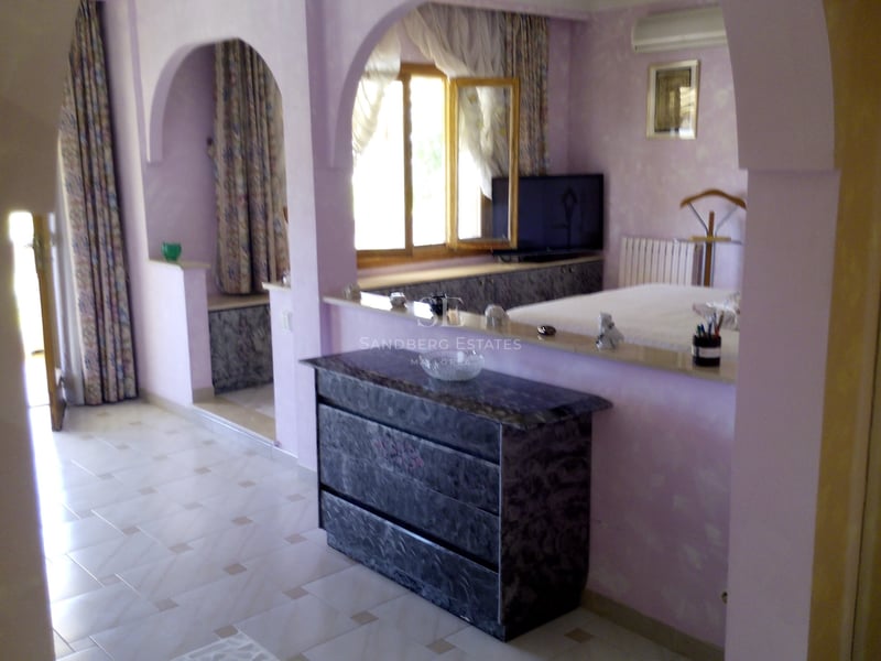 Chambre de maître aux tons lilas avec des arches architecturales, commode en marbre et sol en carrelage clair.