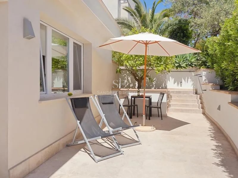 Terrasse en pierre ensoleillée avec deux chaises longues grises, un parasol blanc et un coin repas entouré de verdure.