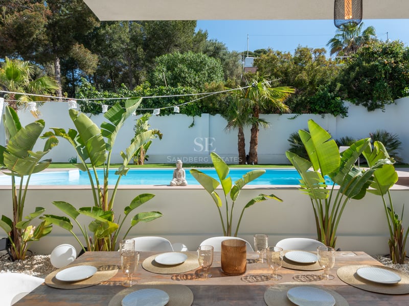 Table de repas en bois devant une piscine turquoise entourée de plantes tropicales et de murs blancs.
