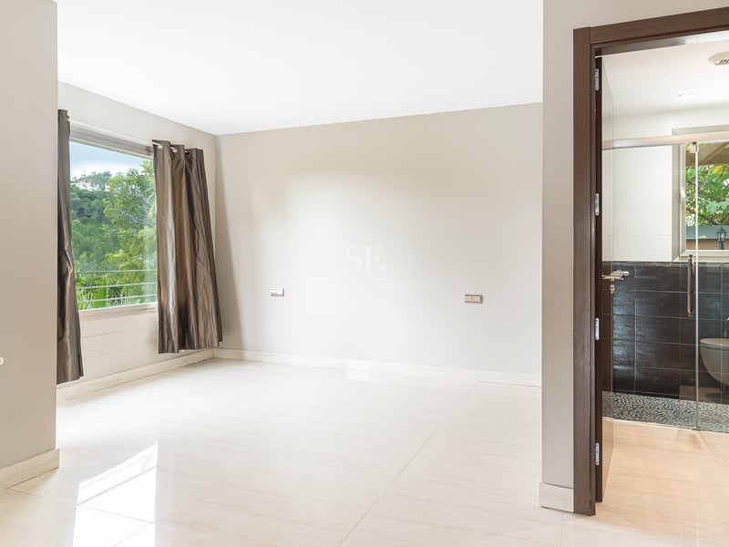 Chambre moderne vide avec sol poli, fenêtre avec rideaux et porte ouverte sur une salle de bain aux tons sombres.