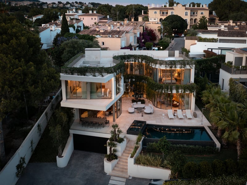Vue aérienne d'une villa contemporaine blanche avec terrasses à niveaux, verdure intégrée et piscine au crépuscule.
