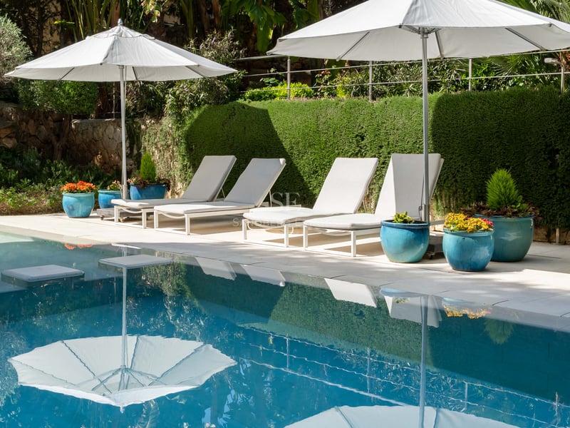 Piscine luxueuse avec quatre chaises longues blanches et parasols assortis devant une haie verte et pots bleus.