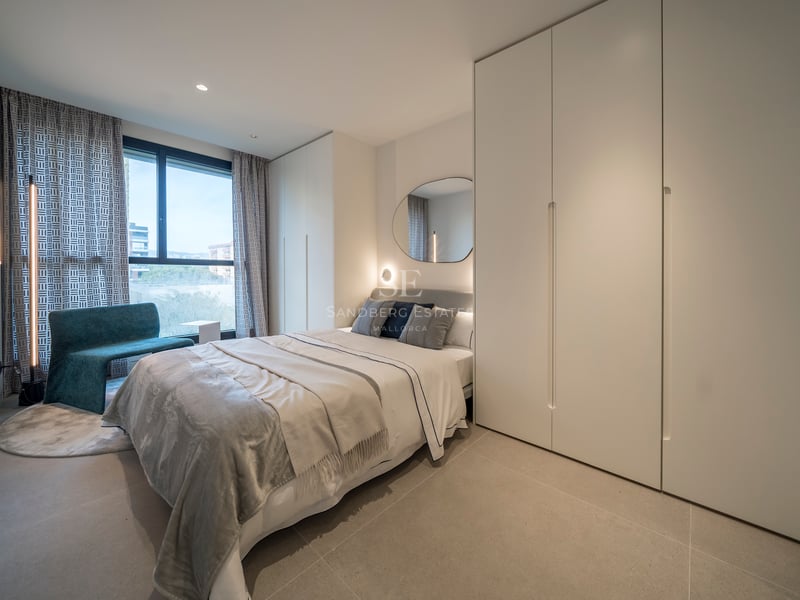 Une chambre moderne lumineuse avec un lit gris, de grandes armoires blanches, une chaise sarcelle et une grande fenêtre.
