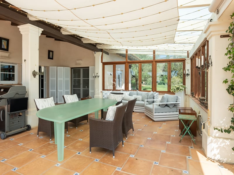 A covered terrace featuring terracotta tiles, a large dining table, a lounge area, and a BBQ grill under a canvas awning.