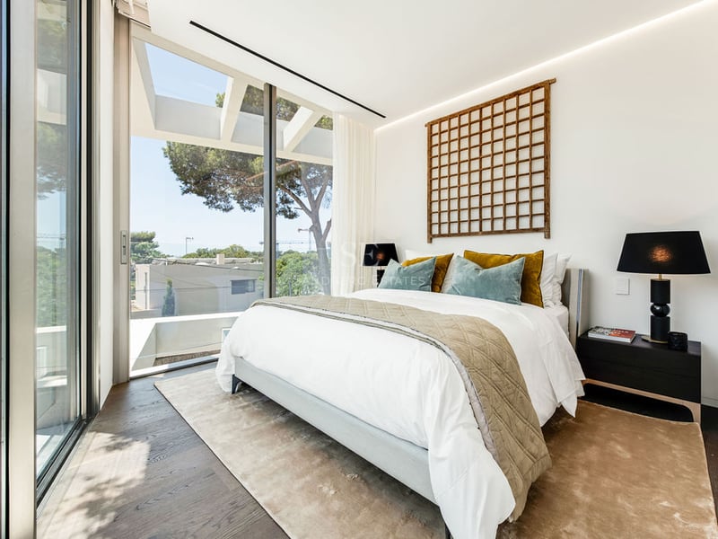 Chambre lumineuse avec parquet foncé, grande baie vitrée et lit blanc orné de coussins en velours bleu et moutarde.