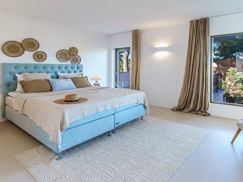 Chambre lumineuse avec lit bleu clair, tapis en laine et grande fenêtre donnant sur des arbres verts.