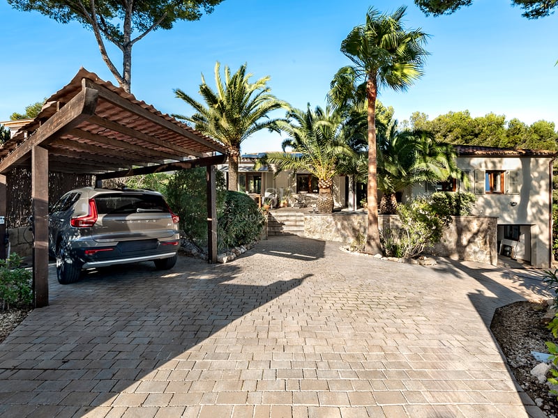 Allée pavée de pierre avec un abri de voiture en bois, des palmiers et une façade de villa en arrière-plan.