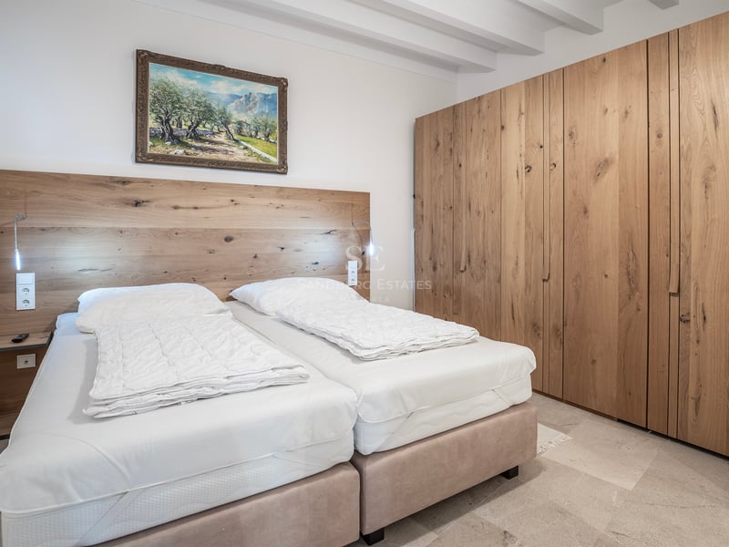 Chambre lumineuse avec plafond à poutres blanches, grand placard en chêne et lits avec tête de lit en bois.