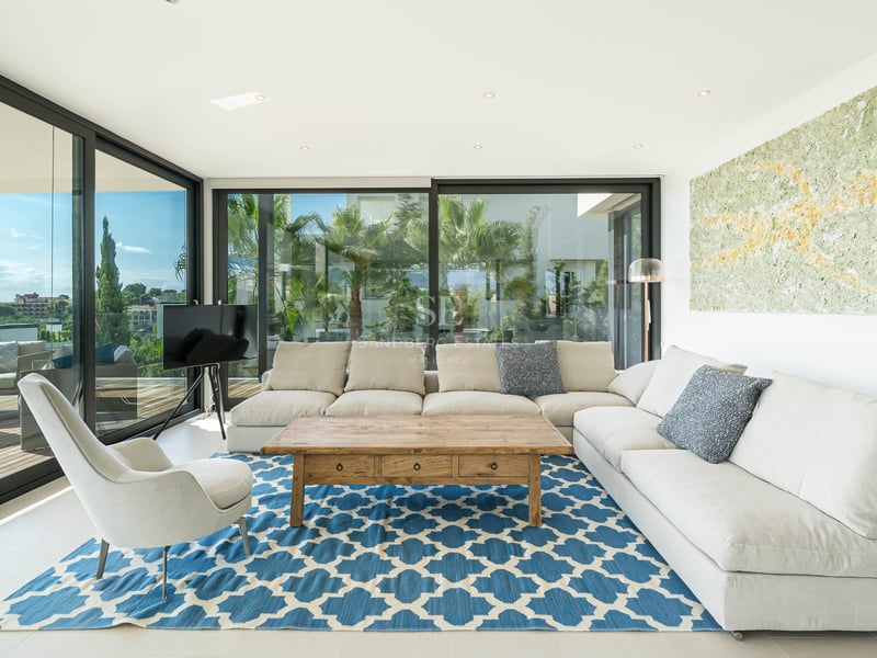 Salon moderne avec canapé en L blanc, tapis bleu à motifs et baies vitrées offrant une vue sur la côte.