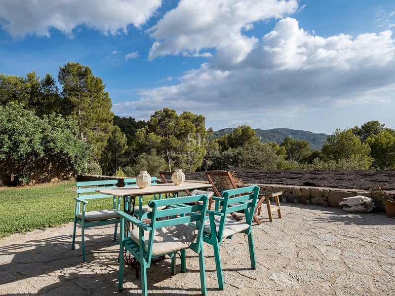 Terrasse en pierre avec table blanche et chaises turquoise entourée de forêts et de montagnes sous un ciel bleu.