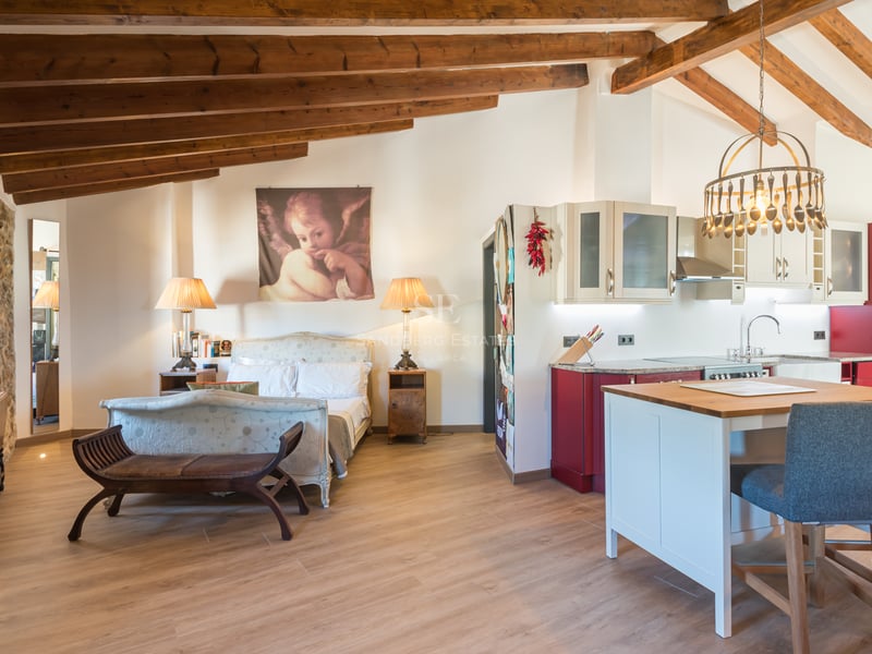 Studio avec lit classique, poutres en bois apparentes, mur en pierre et cuisine rouge et blanche avec îlot.