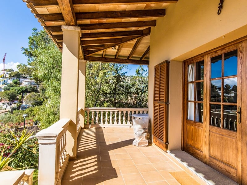 Terrasse couverte avec poutres en bois, sol carrelé et balustrade en pierre surplombant un paysage verdoyant.