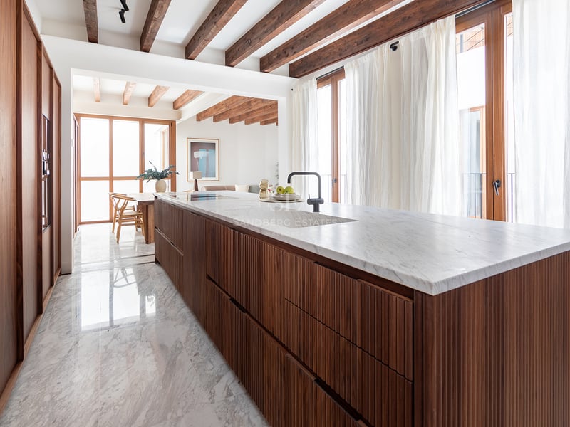 Cocina moderna con isla de madera alistonada, encimera de mármol blanco, suelos de piedra y vigas de madera en el techo.
