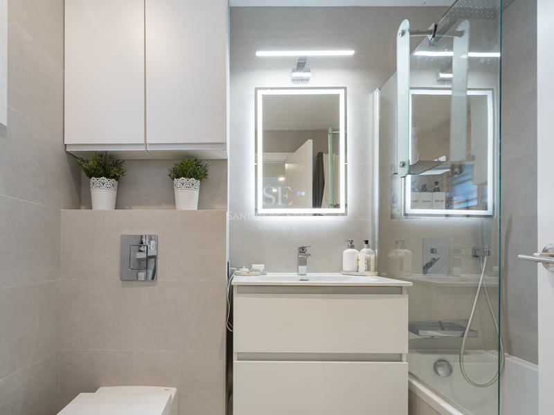 Salle de bain moderne avec vanité blanche, miroir LED, paroi en verre et carrelage beige grand format.