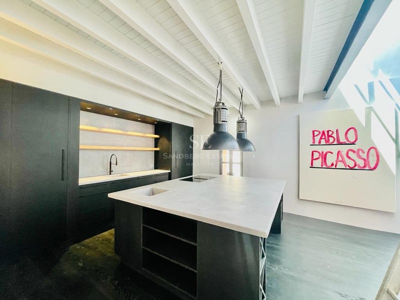 Cuisine moderne avec îlot central sombre, plan de travail gris et plafond à poutres blanches.