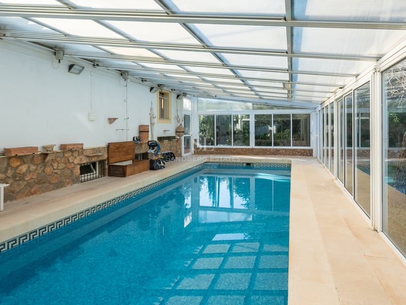Piscine intérieure avec mosaïques bleues, murs en pierre et toit translucide à côté de grandes fenêtres.