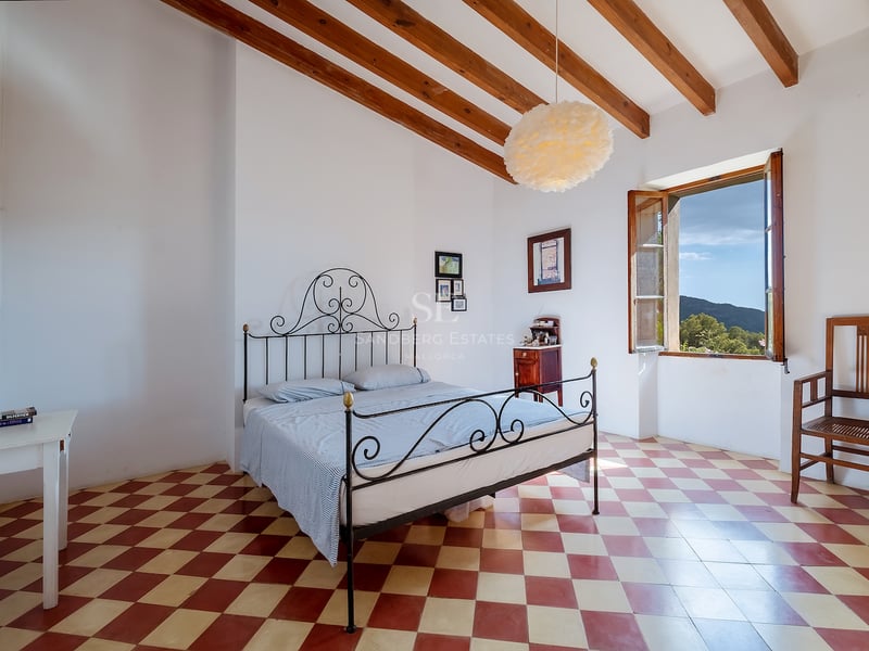 Chambre avec lit en fer forgé, sol en damier rouge et blanc, poutres en bois et fenêtre donnant sur les collines.