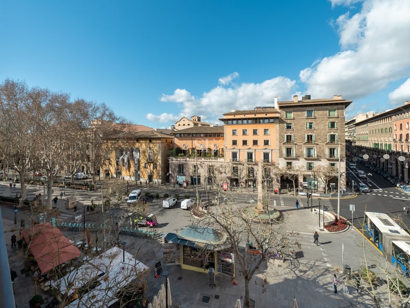 High-angle view from a wrought iron balcony overlooking a bustling historic city plaza with sandstone buildings and trees.