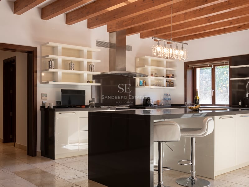 Cuisine moderne avec îlot noir, placards blancs, poutres en bois apparentes et sol en pierre naturelle.