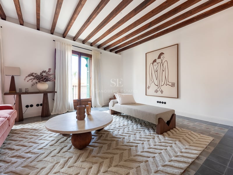 Salón luminoso con vigas de madera oscura, alfombra de espiga, mesa de travertino y acceso al balcón.