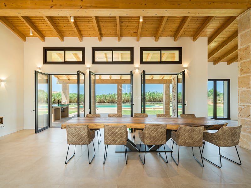 Comedor espacioso con mesa de madera natural, techo de vigas vistas y pilar de piedra con vistas a la piscina.