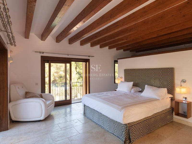 Chambre de maître avec poutres en bois au plafond, sol en pierre calcaire et accès au balcon.