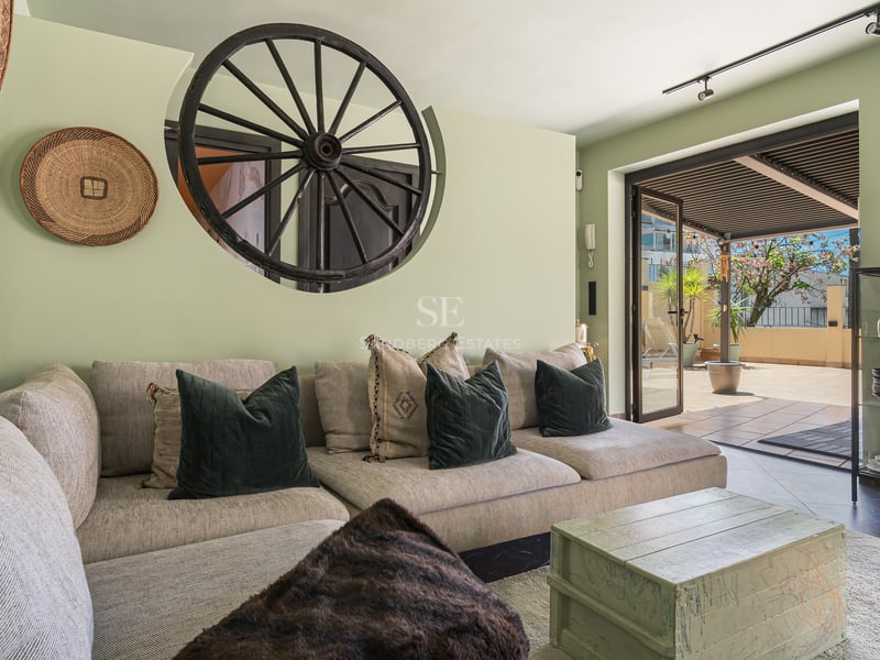 Salon chaleureux avec canapé beige, mur vert sauge et accès direct à une terrasse ensoleillée sous pergola.