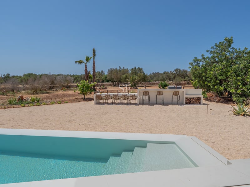 Modern white swimming pool foreground with outdoor dining area and bar set against a Mediterranean landscape.
