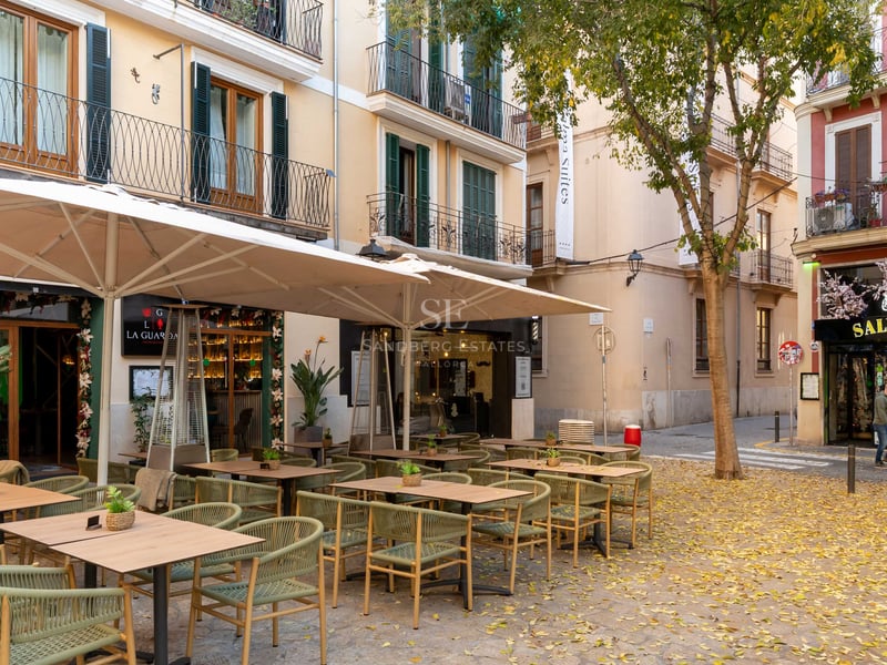 Outdoor restaurant terrace with sage green chairs under cream umbrellas in a historic Mediterranean city street.