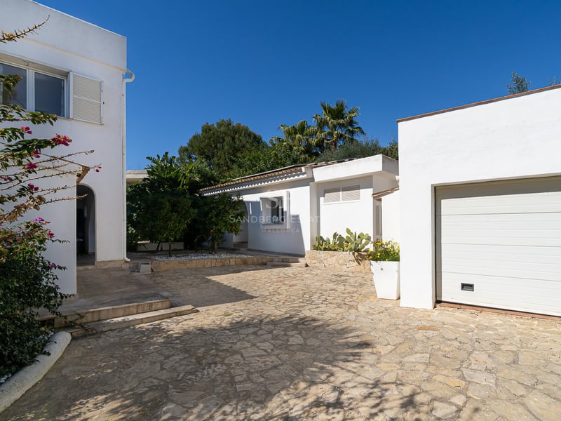 Villa méditerranéenne blanche avec une allée pavée en pierre, un garage et une végétation luxuriante sous un ciel bleu.
