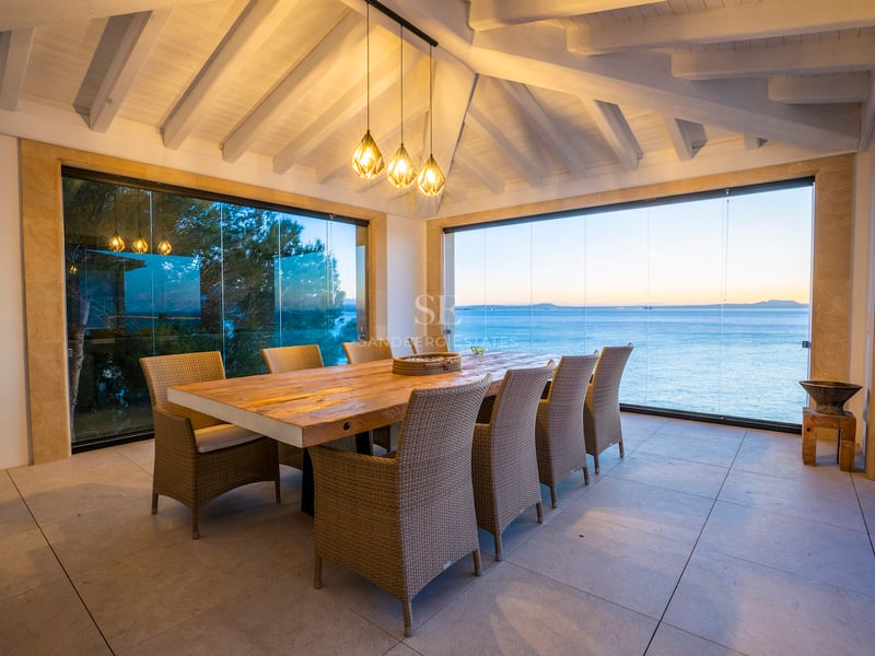 Grande table à manger en bois sur une terrasse avec rideaux de verre surplombant la mer au crépuscule.