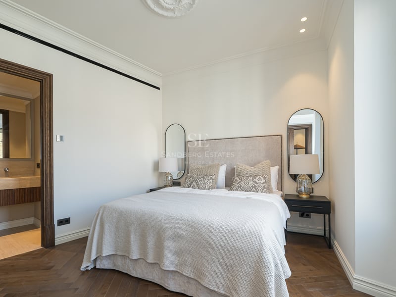 Luxury bedroom featuring oak herringbone floors, a large bed, and a view into a travertine marble en-suite bathroom.