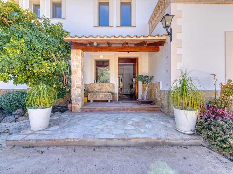 Entrée de villa avec piliers en pierre, porche en bois, murs blancs et plantes en pots sous une lumière naturelle.