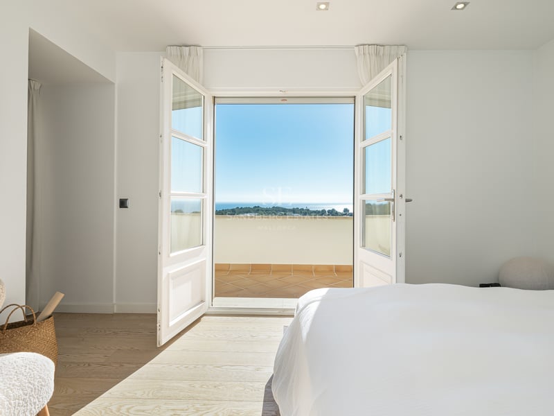 Chambre blanche lumineuse avec portes-fenêtres ouvertes sur une terrasse ensoleillée surplombant la mer.