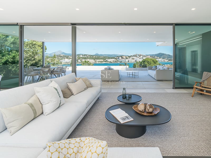 Salon blanc moderne avec baies vitrées et terrasse offrant une vue panoramique sur la mer et les montagnes.
